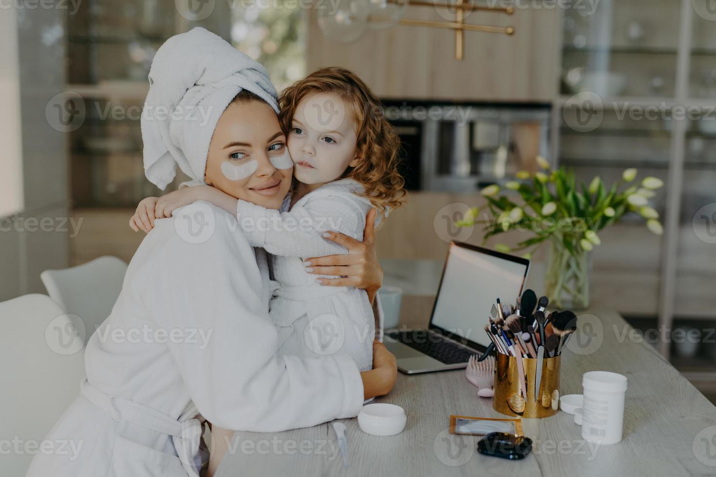 Attractive woman wears hydrogel patches under eyes dressed in bathrobe and wrapped towel embraces her small daughter with love pose together at home near cosmetic tools. Makeup routine concept photo