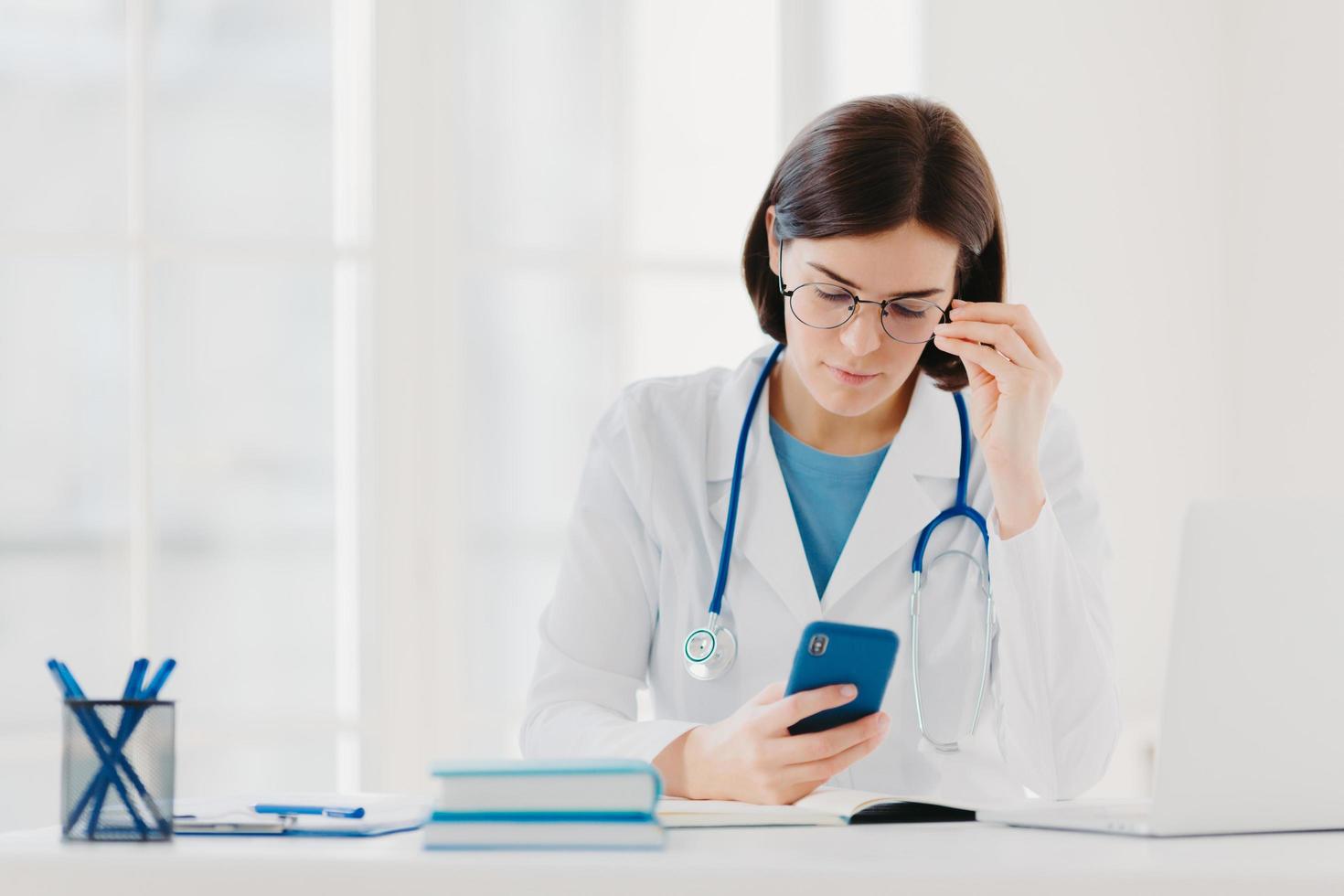 Medical technology concept. Concentrated woman doctor looks at smartphone device, checks necessary information in internet, wears stethoscope around neck, big round glasses, poses in clinic. photo