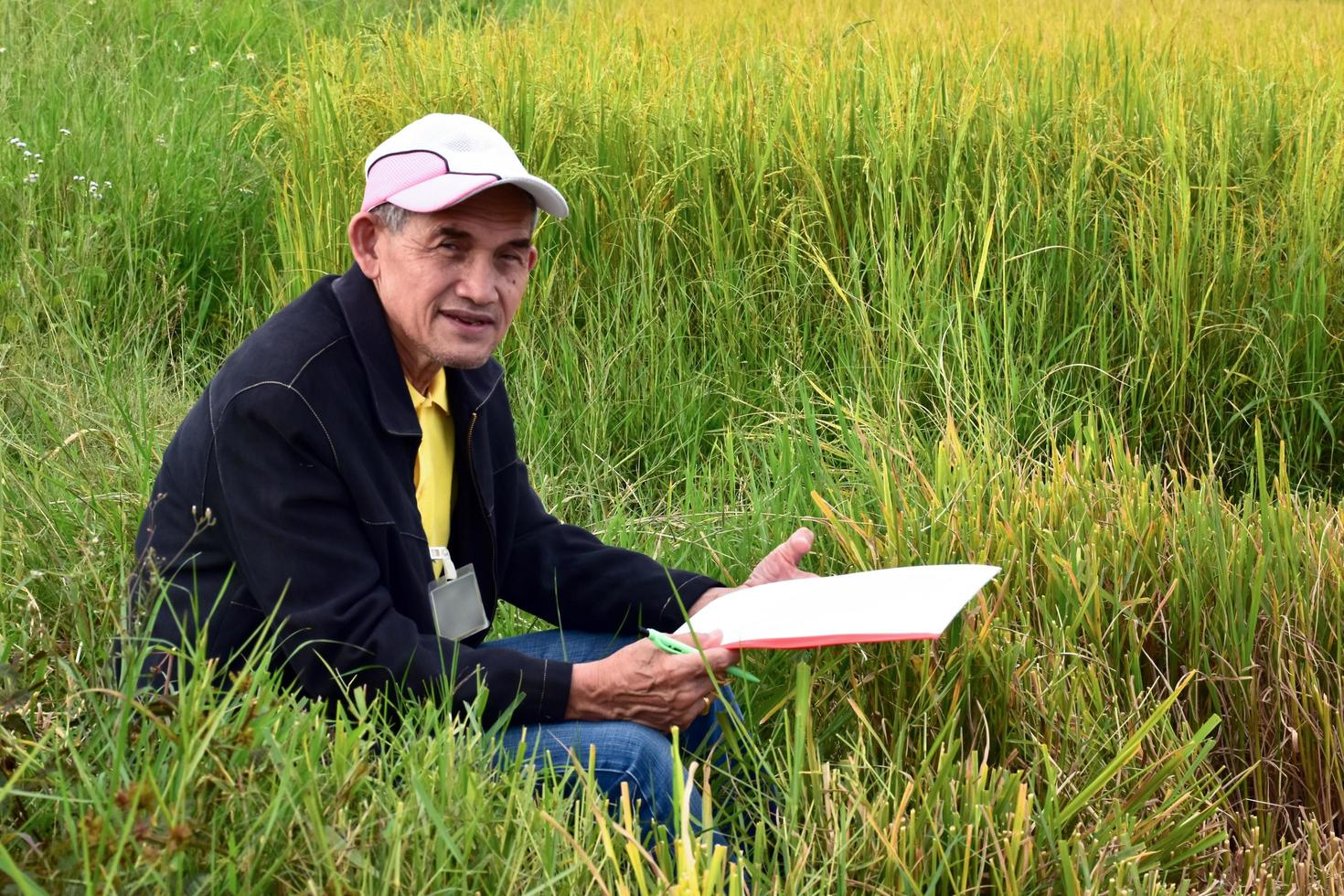 retrato de un voluntario senior asiático que tiene papel blanco y bolígrafo en la mano, se sienta en la hierba cerca del campo de arroz, concepto de vida feliz después de la jubilación de personas asiáticas amables y serviciales. foto