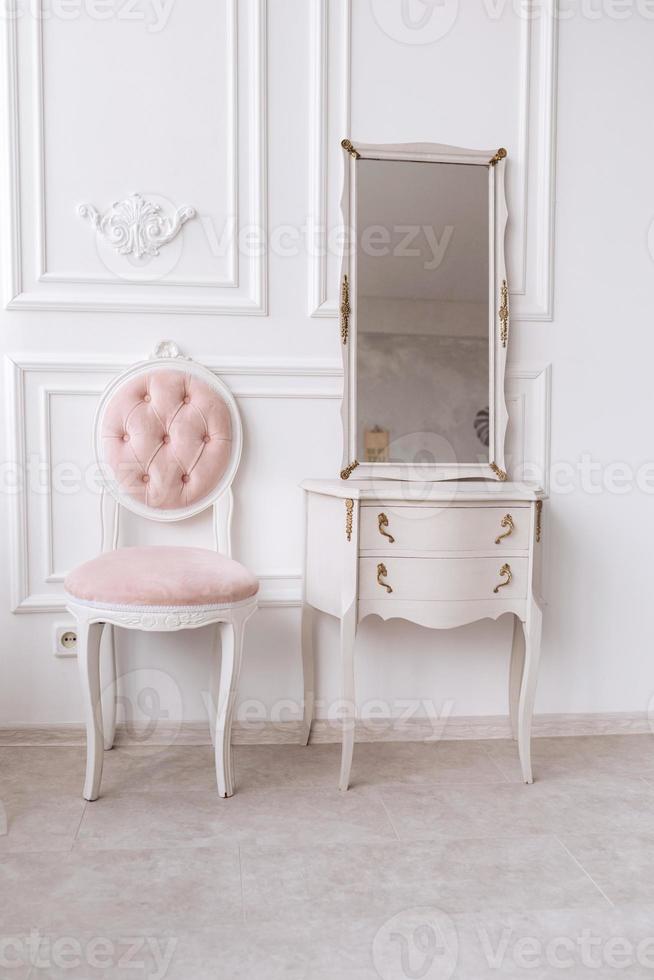 classic room with white patterned wall, dressing table with mirror, decorated with golden and pink soft chair. Details of the interior of the bedroom for girls and make-up, hairstyles with a mirror photo