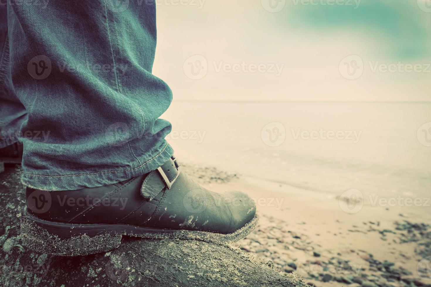 Man in jeans and elegant shoes standing on fallen tree on wild beach looking at sea. Vintage photo