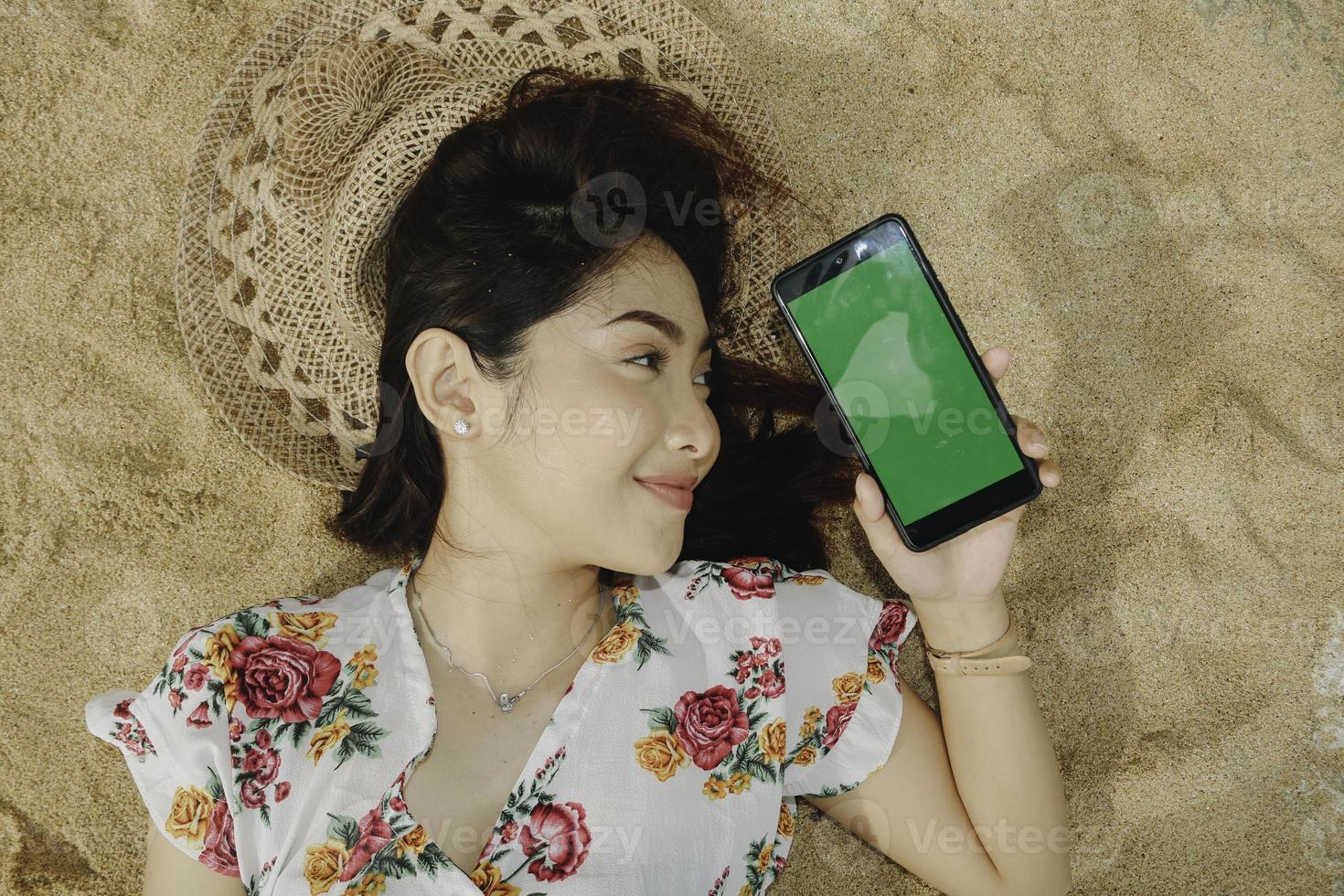 A young beautiful Asian woman lying on the sand while showing phone screen photo
