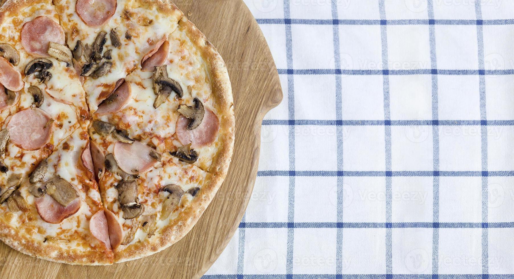 pizza con jamón y champiñones en una tabla de madera y servilleta azul, vista superior. foodphoto para pizzería, menú, catálogo, pancarta, folleto foto