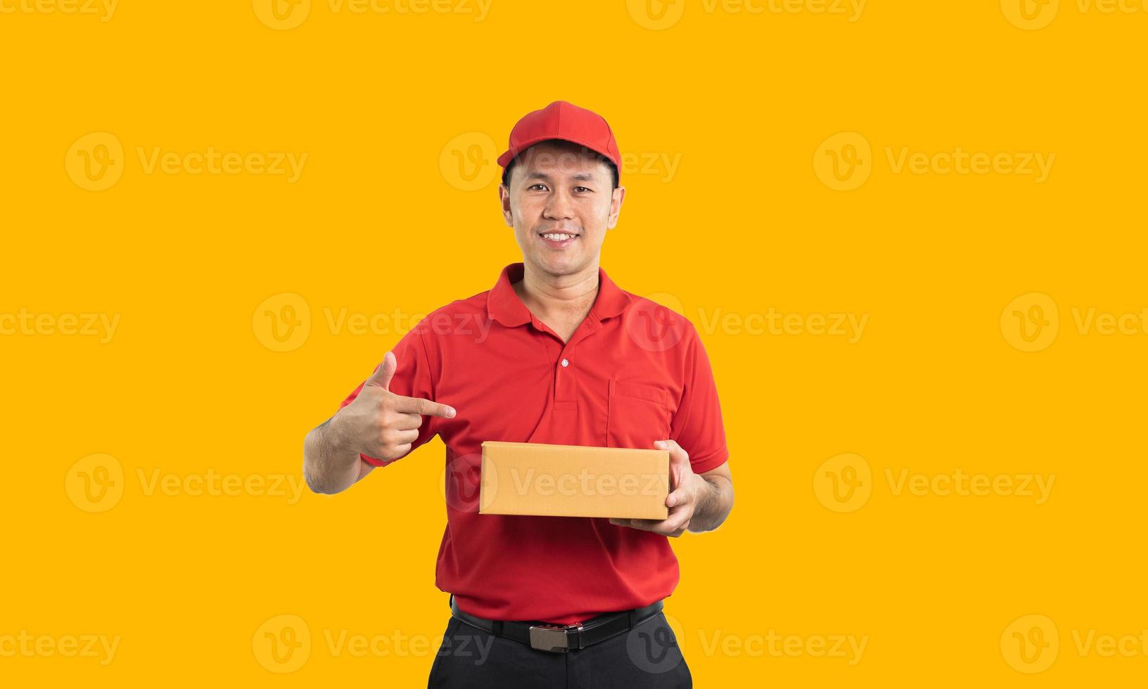 Asian delivery man worker smiling and pointing finger at the box in red uniform isolated on yellow background, hold parcel boxes for sending or conveying parcels by mail. photo