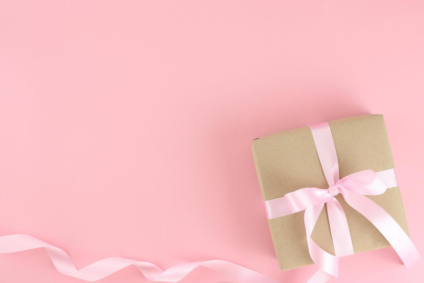 Brown paper gift box with pink satin curly ribbon bow on pastel pink background. Flat lay mother day, father day, valentines day, birthday concepts with copy space. photo