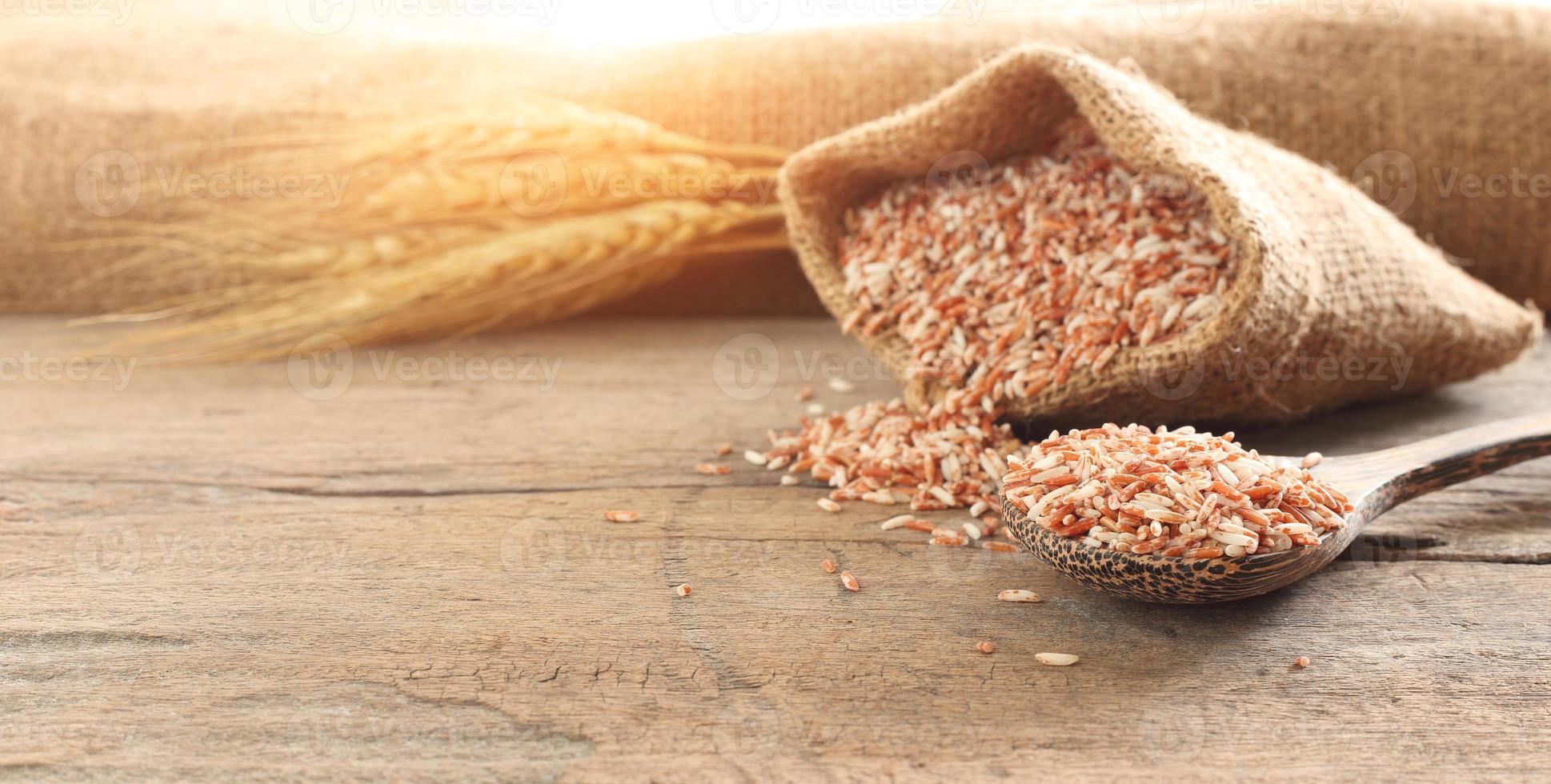 Healthy food - Close up spoon of red organic whole rice grain and brown sack on wooden table with ear  of paddy against shiny background photo