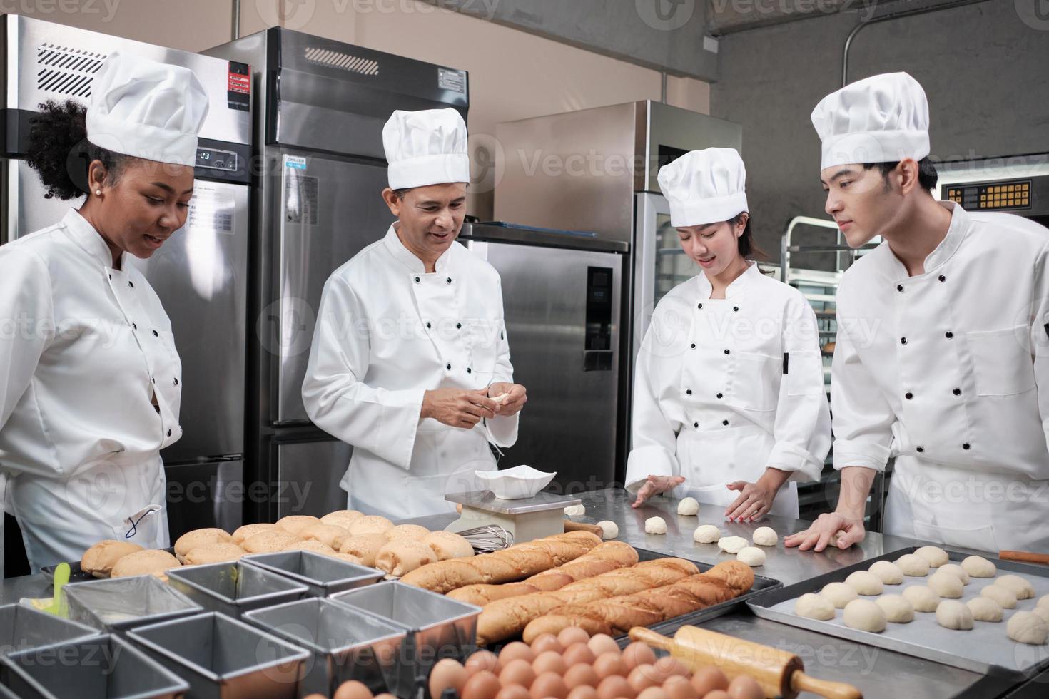 Multiracial professional gourmet team, four chefs in white cook uniforms and aprons knead pastry dough and flour, prepare bread, and bakery food, baking in oven at stainless steel restaurant kitchen. photo