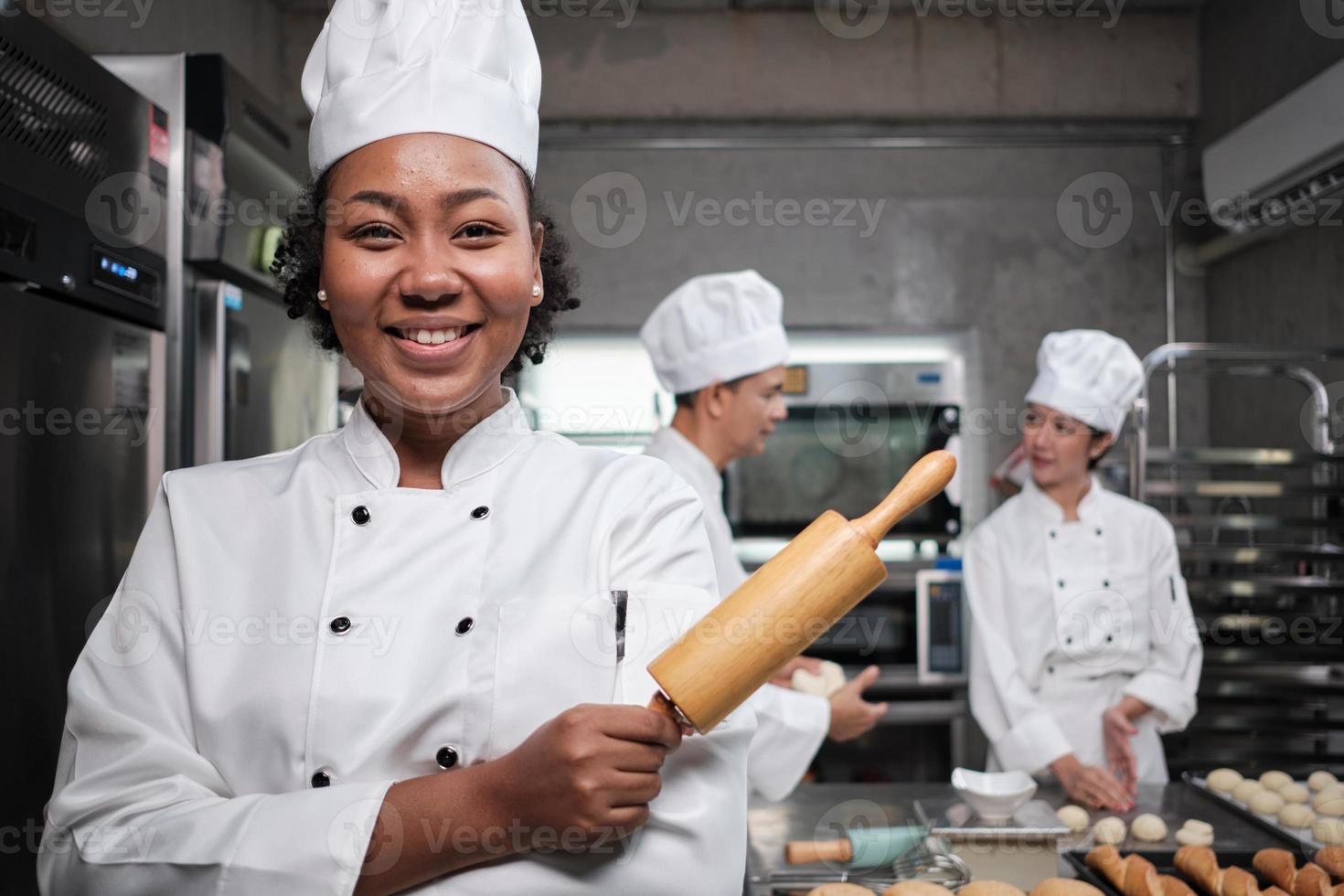 Portrait of young African American female chef in white cooking uniform looking at camera, cheerful smile with foods professional occupation, commercial pastry culinary jobs in a restaurant kitchen. photo