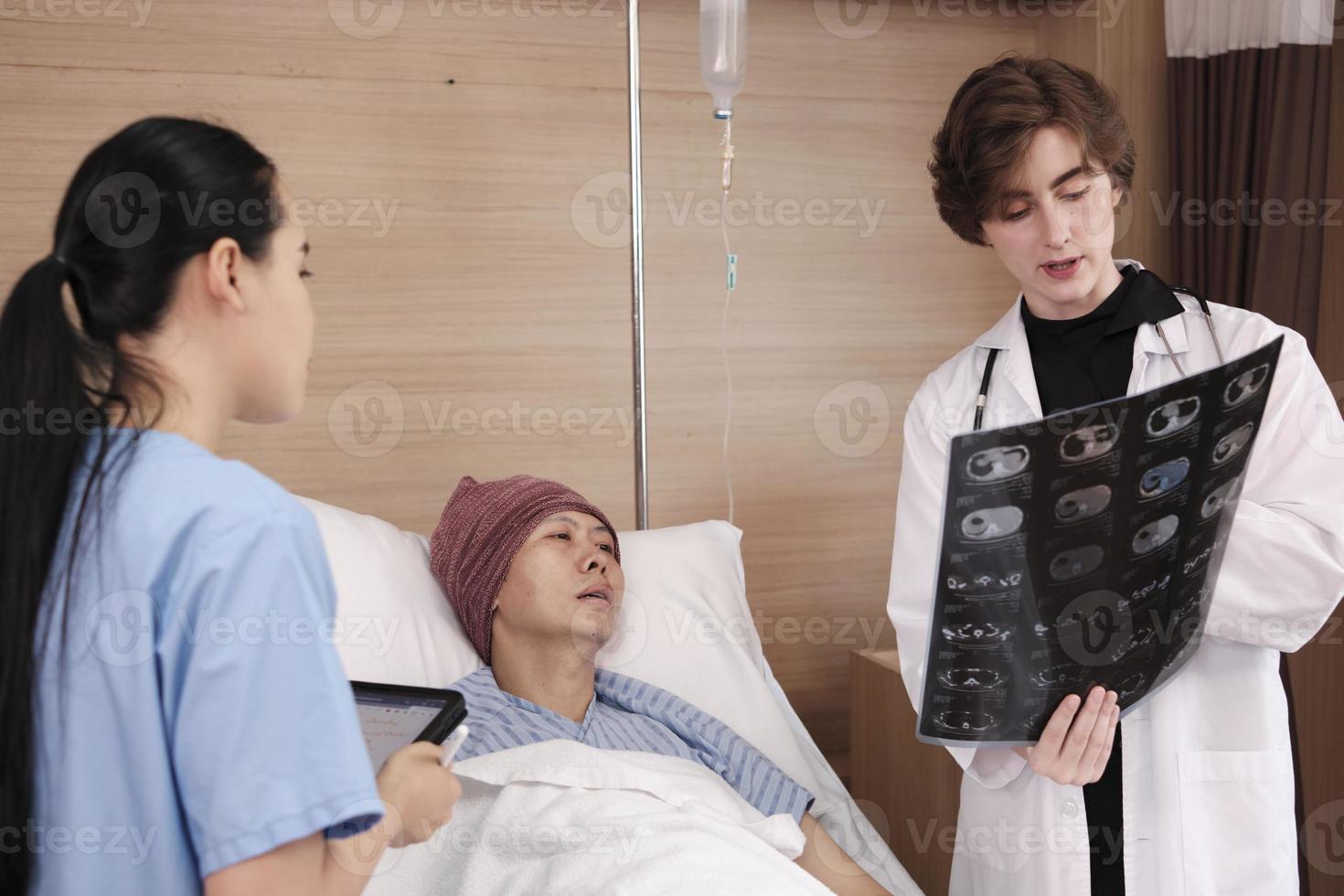 Caucasian female doctor in uniform diagnosis explains x-ray film with Asian radiologist and recovery male patient at inpatient room bed in a hospital ward, medical clinic, cancer examination consult. photo