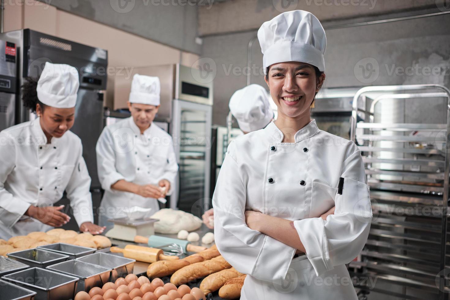 Young beautiful Asian female chef in white cooking uniform looks at camera, arms crossed and cheerful smile with food professional occupation, commercial pastry culinary jobs in a restaurant kitchen. photo