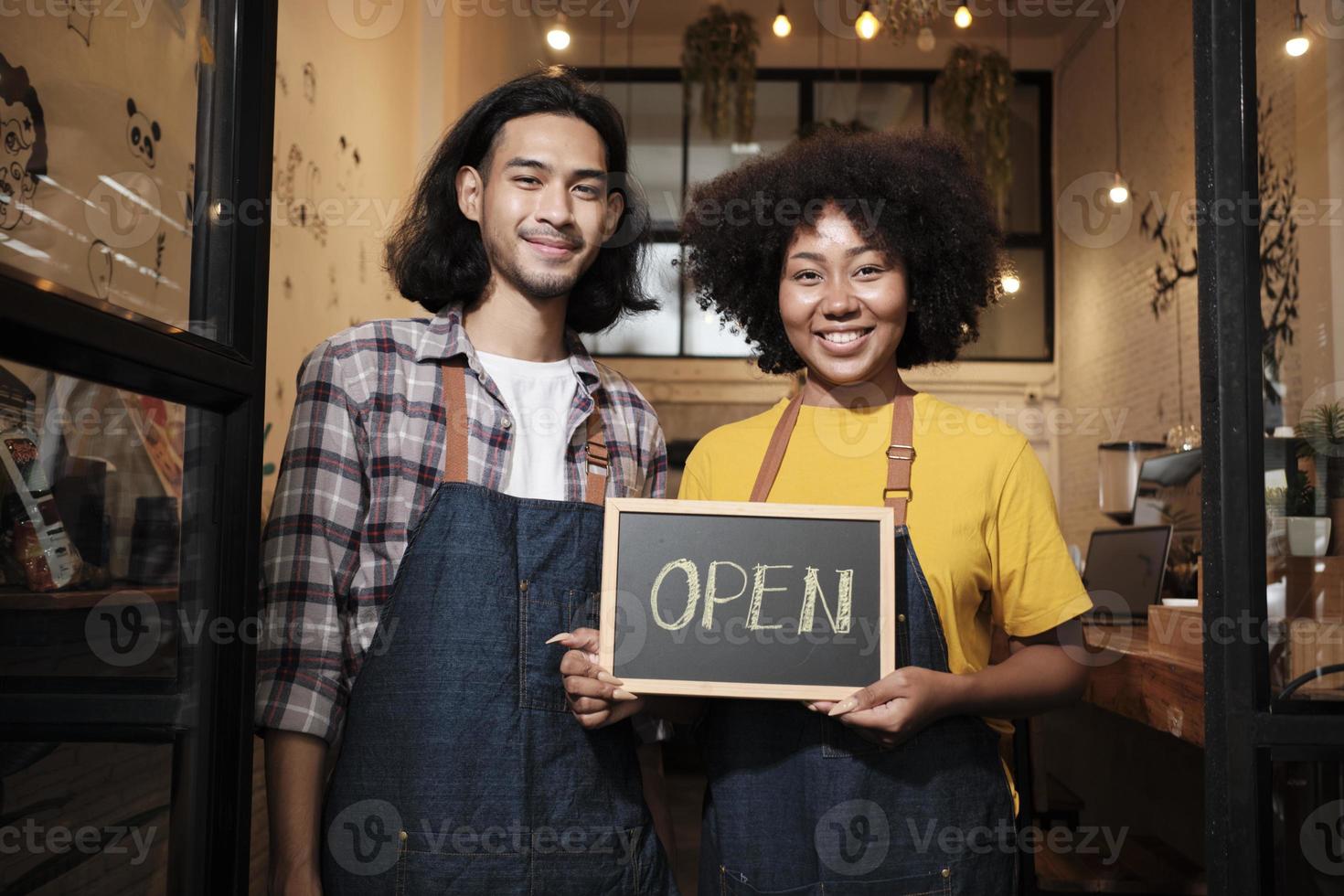 Two young startup barista partners with aprons stand at casual cafe door, letters on board and show open sign, happy and cheerful smiles with coffee shop service jobs, and new business entrepreneurs. photo