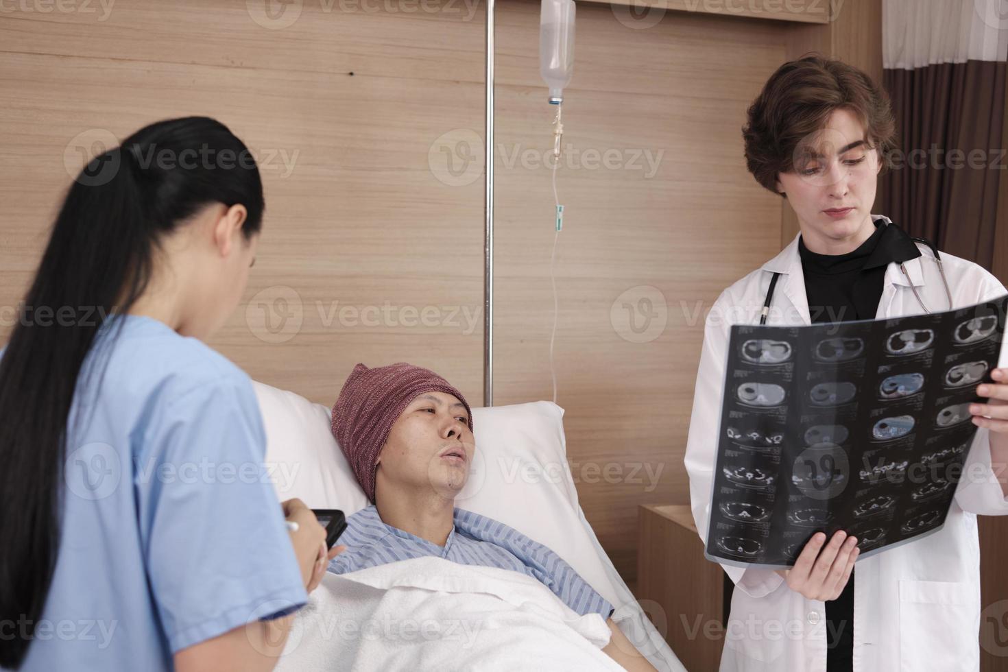 una doctora caucásica con diagnóstico uniforme explica la película de rayos X con un radiólogo asiático y un paciente varón en recuperación en la cama de una habitación para pacientes hospitalizados en una sala de hospital, clínica médica, consulta de examen de cáncer. foto