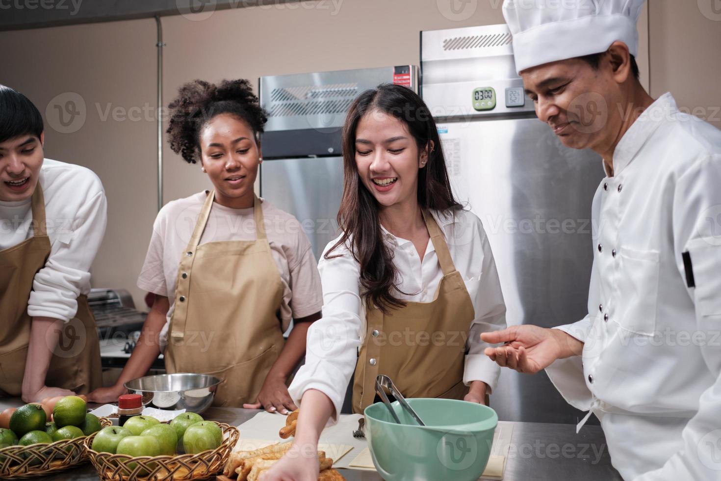 Cuisine course, senior male chef in cook uniform teaches young cooking class students to knead and roll pastry dough, prepare ingredients for bakery foods, fruit pies in stainless steel kitchen. photo