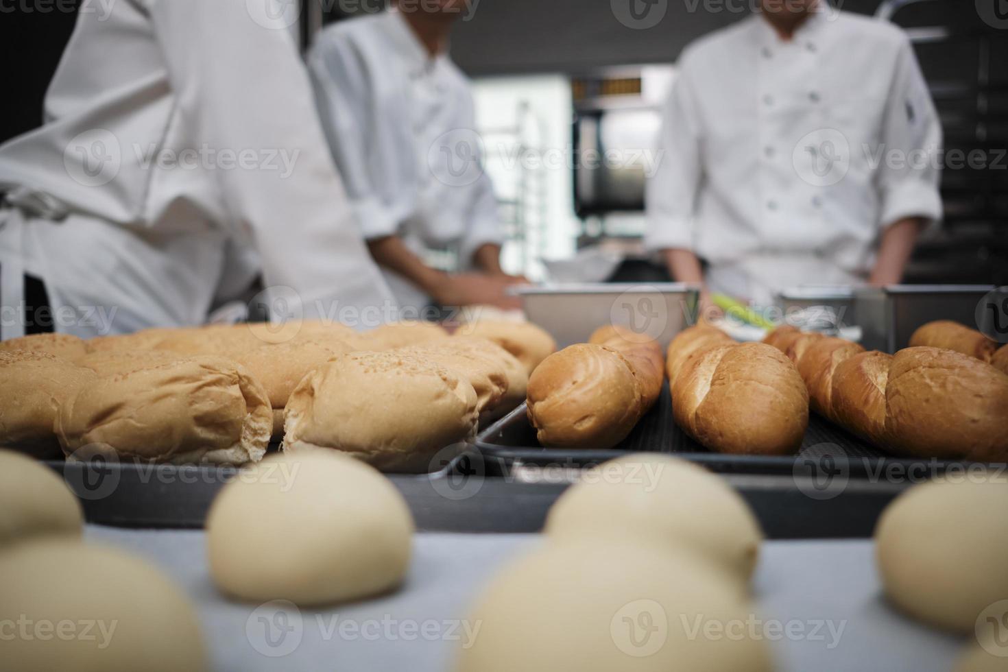 Close-up shot and selective focus at baguettes and delicious bread in front of white uniform chefs team, kneading raw pastry dough, preparing fresh bakery food, baking in oven in restaurant kitchen. photo