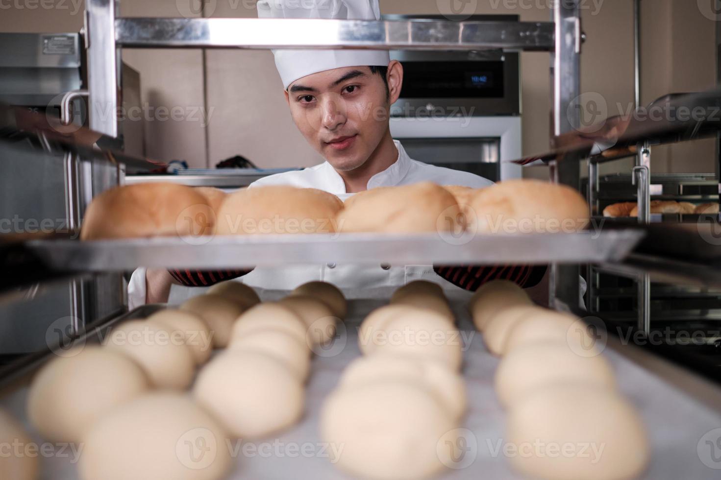 Young professional Asian male chef in white cook uniform with hat, gloves, and apron making bread from pastry dough, preparing fresh bakery food, baking in oven at restaurant stainless steel kitchen. photo