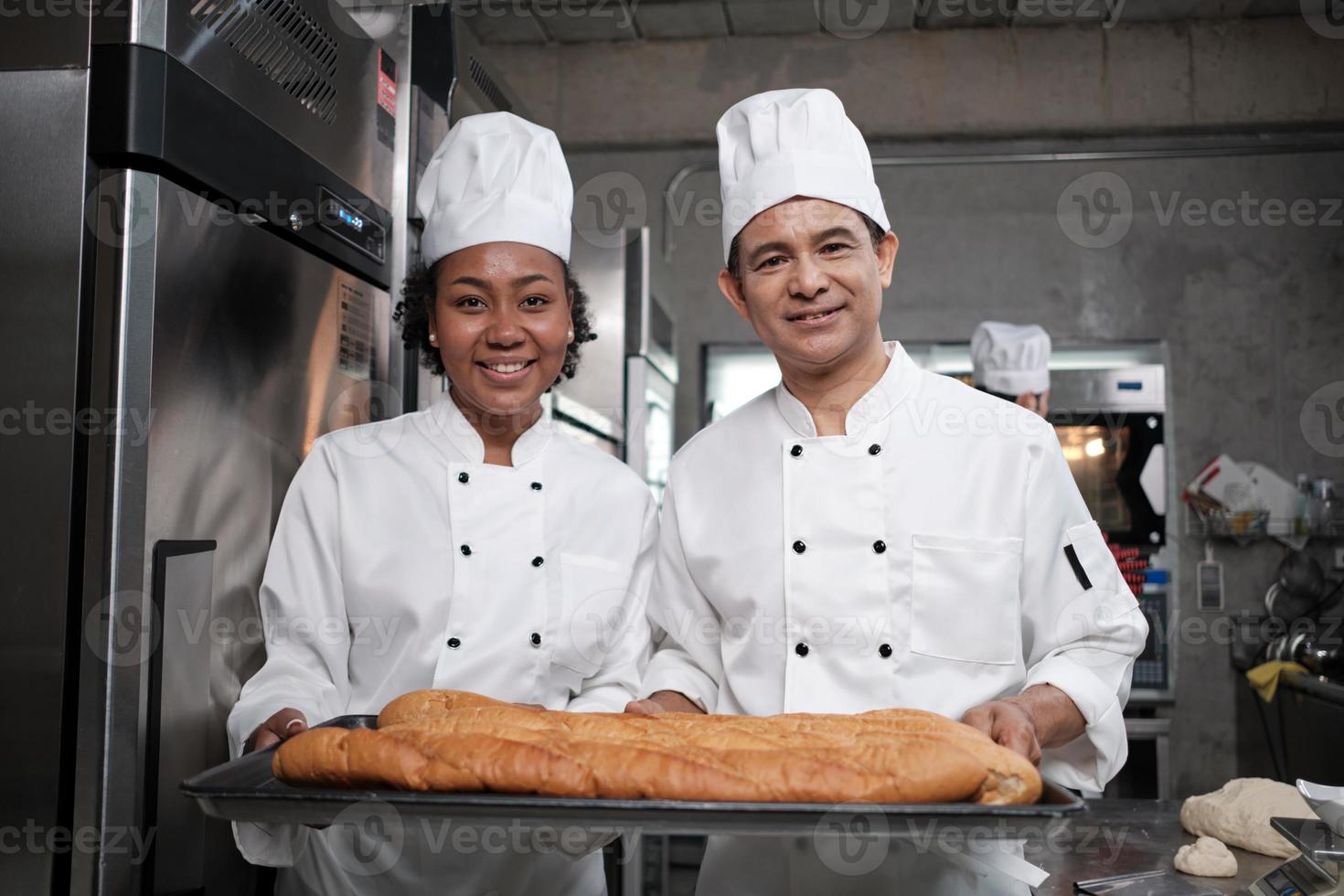Portrait of professional chefs in white uniform looking at camera with cheerful smile and proud with tray of baguette in kitchen. Friend and partner of pastry foods and fresh daily bakery occupation. photo
