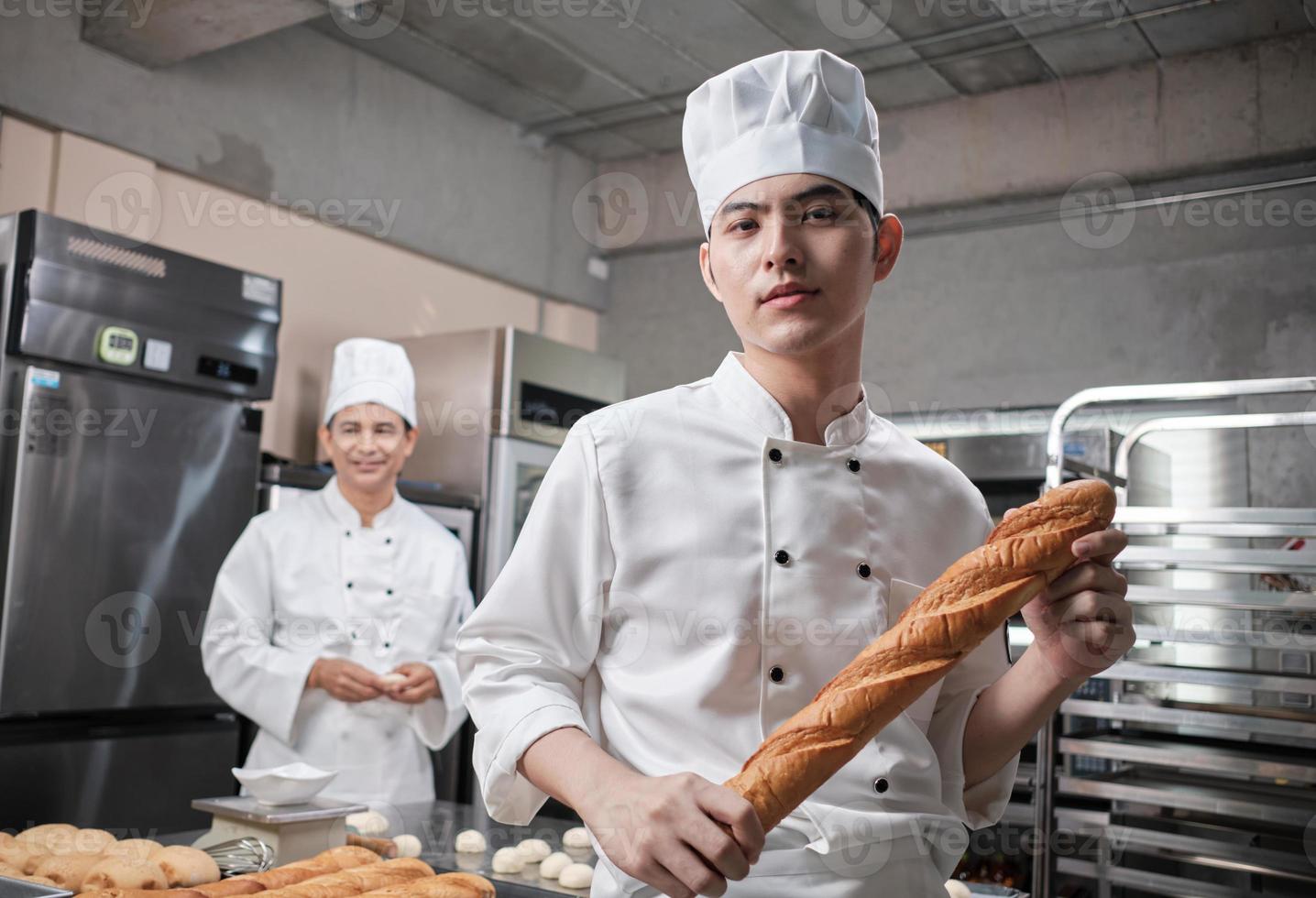 Portrait of young Asian male chef in white cooking uniform looks camera, happy smile and cheerful with baguette, professional foods occupation, commercial pastry culinary jobs in a restaurant kitchen. photo