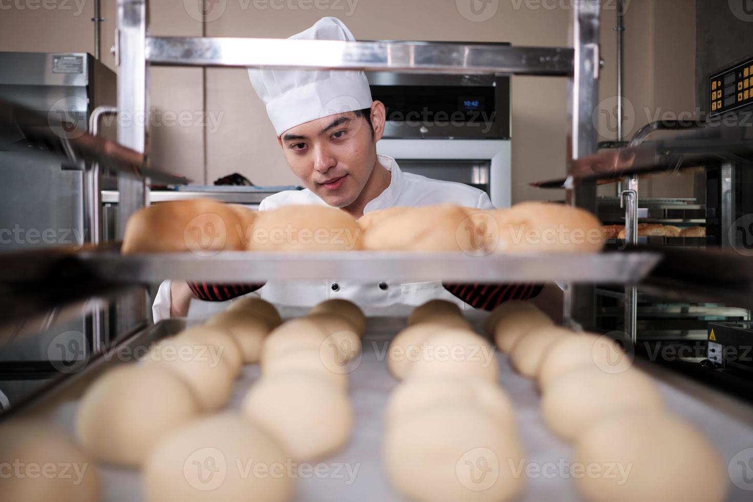 joven chef asiático profesional con uniforme de cocinero blanco con sombrero, guantes y delantal haciendo pan con masa de pastelería, preparando comida fresca de panadería, horneando en el horno en la cocina de acero inoxidable del restaurante. foto