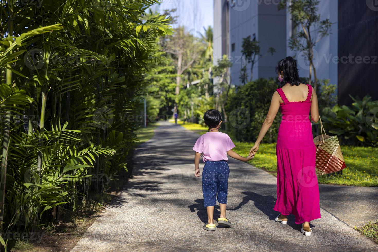 niño asiático y mamá caminan de la mano usando una máscara blanca autoprotectora tomando el sol durante el brote de virus. coronavirus, covid-19, concepto de pandemia, epidemia de covid foto