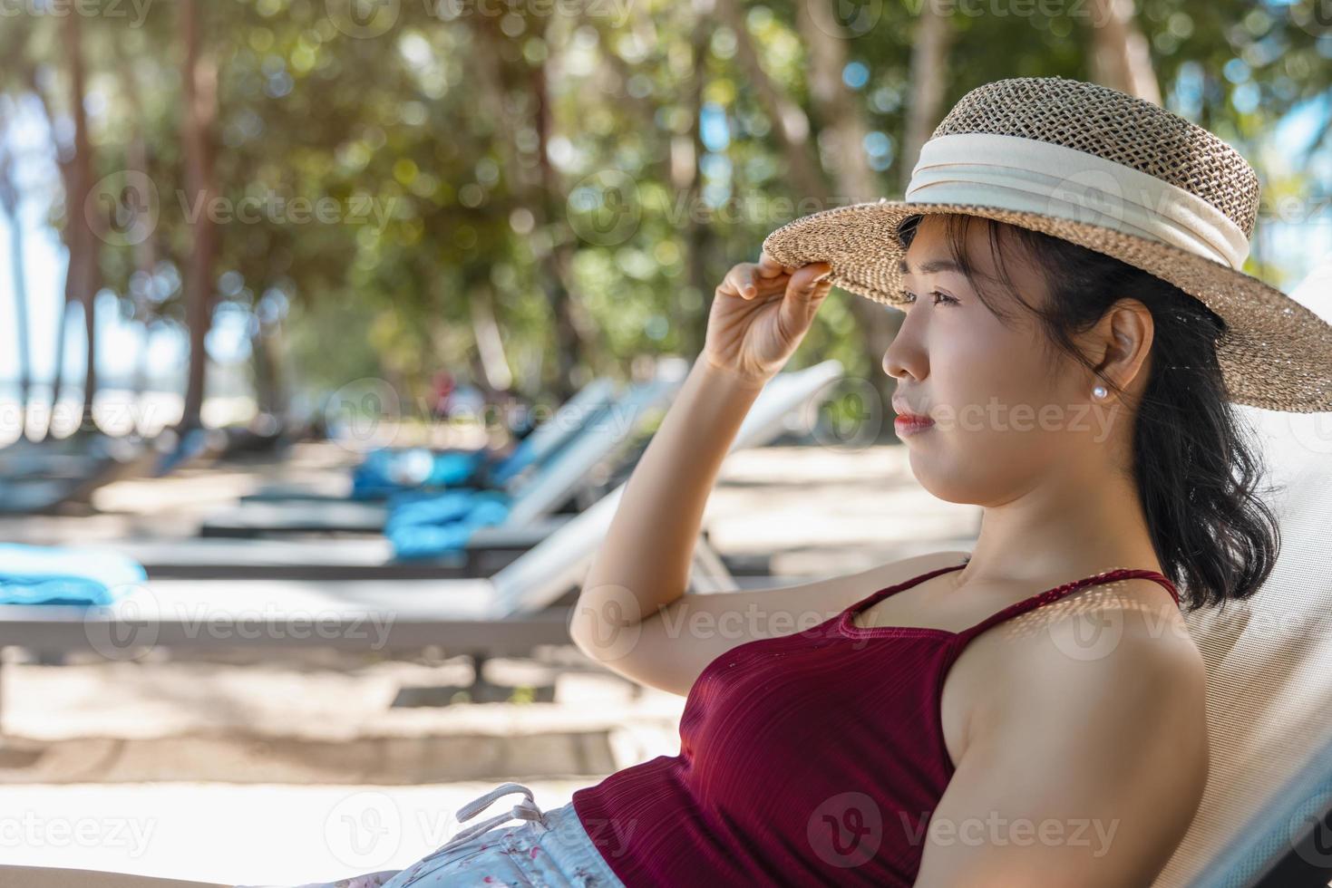 Model Asian woman wearing red swimsuit and straw hat sits in sun by the sea. Beautiful girl relaxing at infinity luxury resort. Spa, wellness photo