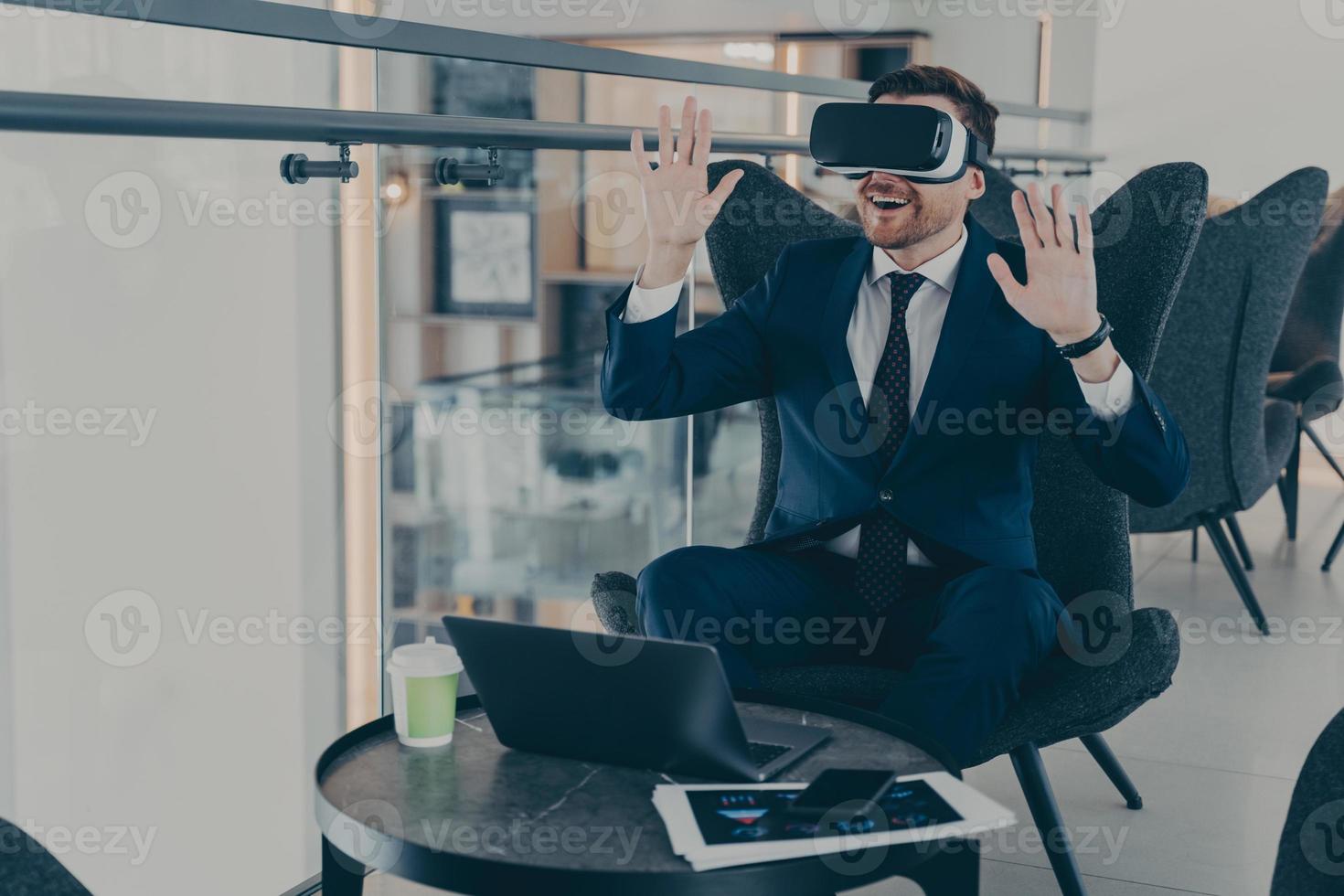 Businessman dressed in formal suit wearing VR goggles while working on laptop in cafe photo