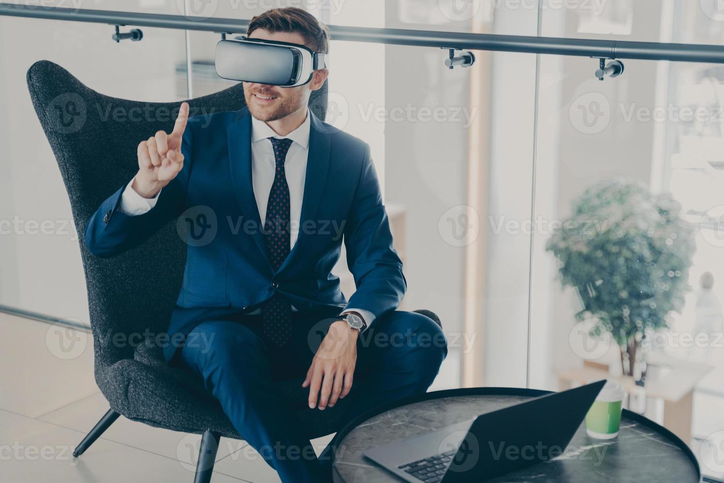 Smiling businessman using VR headset glasses while sitting in office lobby and working on laptop photo