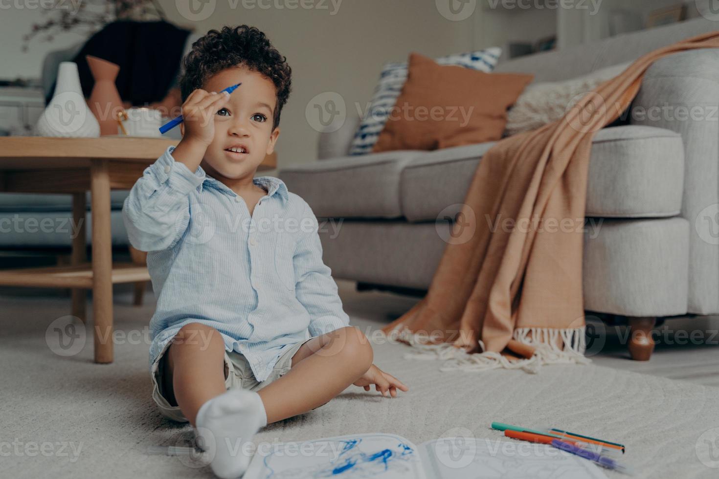 Curious mixed race toddler in casual clothes and white socks sitting on floor while drawing in album photo