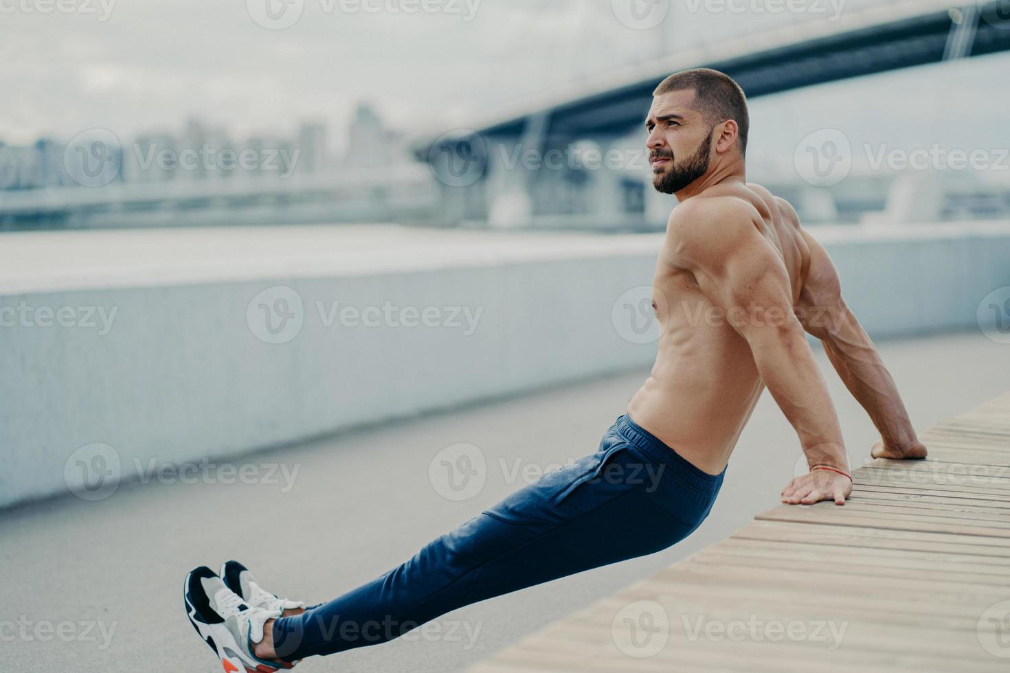 Purposeful bearded adult man trains biceps while doing reverse push up exercises poses with naked muscular torso concentrated away breathes fresh air, likes fitness and sport. Workout outside photo