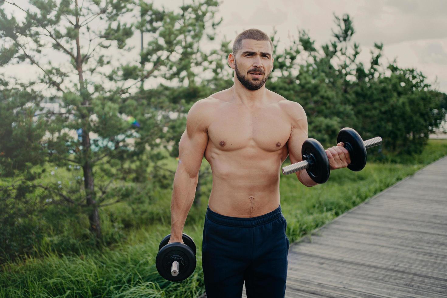 Handsome muscular man lifts barbells outside, does biceps training, stands with naked sexy torso, trains muscles, dressed in shorts poses outdoor, has attractive body. Weight lifting concept photo