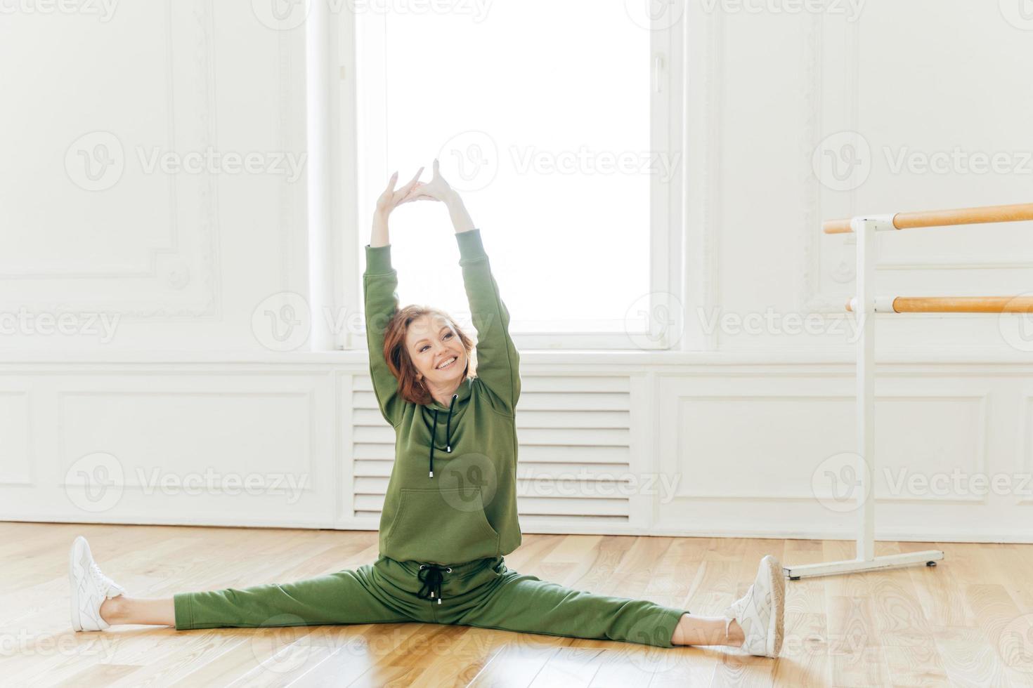 People, strength, fitness, gymnastics concept. Glad sportswoman with cheerful face, raises hands, spreads legs and does splits, stretches in studio. Young dancer has work out, dressed in tracksuit photo