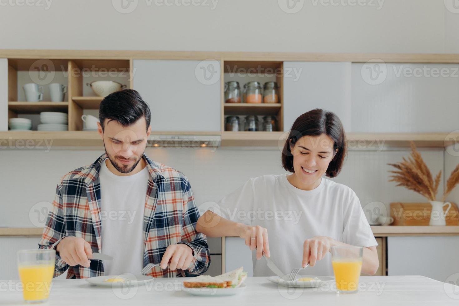 la pareja casada de la familia posa en la mesa de la cocina, toma un desayuno delicioso, habla sobre los planes del día, come huevos fritos y hamburguesas, bebe jugo de manzana fresco, se viste informalmente, disfruta del ambiente doméstico foto