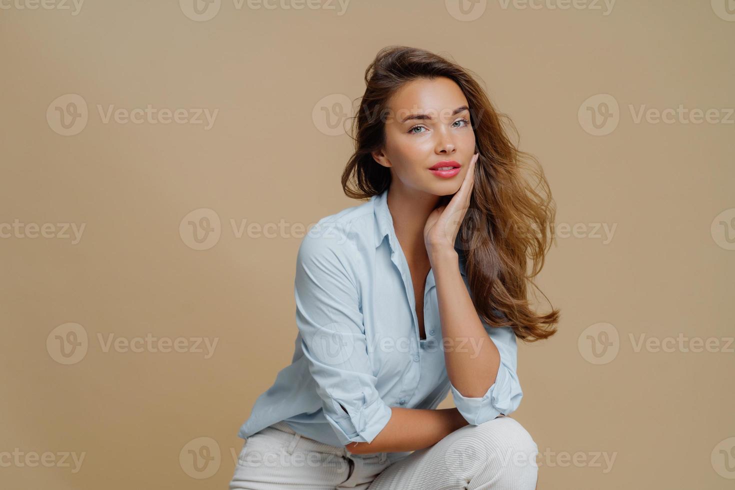 Portrait of attractive young woman sits against brown background, touches cheek, dressed in fashionbale outfit, has thoughtful look at camera, demonstrates her beauty. People and style concept photo