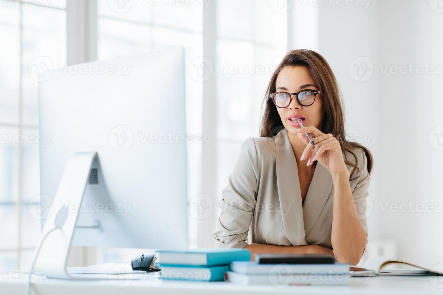 Contemplative female entrepreneur keeps pen in mouth, focused in monitor of computer, thinks on development of new strategy, wears elegant clothes and spectacles, poses against office interior photo