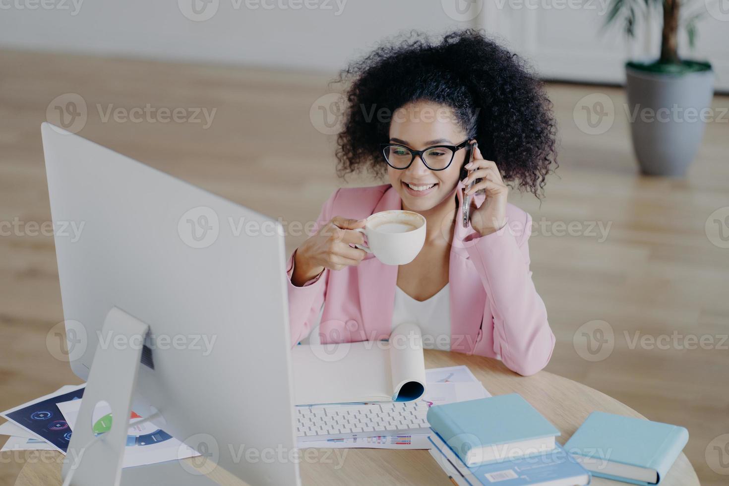 Glad dark skinned business lady looks happily at computer, drinks fresh hot beverage, holds modern mobile phone, dressed elegantly, poses against office interior, sits at desk with textbook, notepad photo