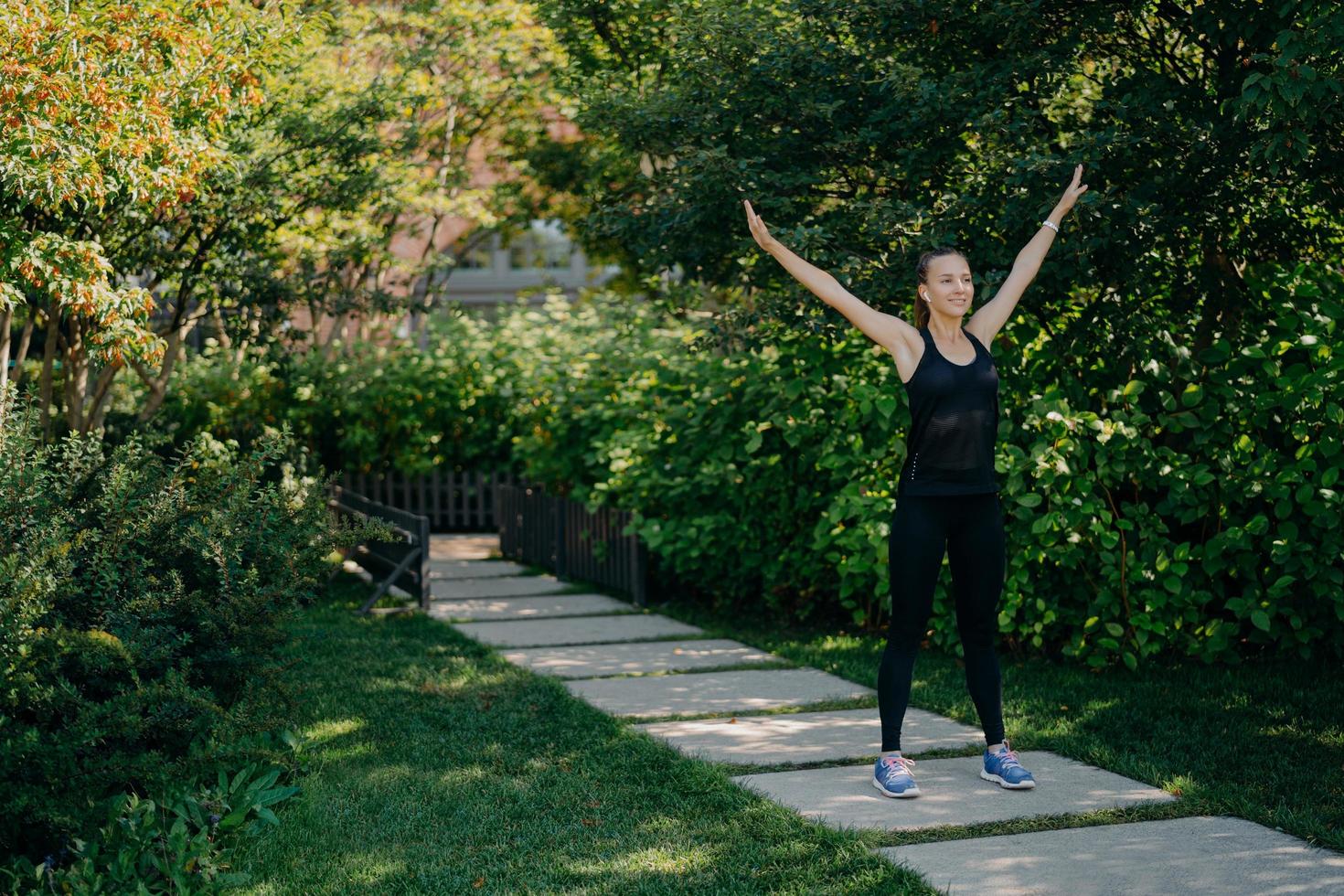 Full length shot of active sportswoman raises arms breathes deeply dressed in active wear has sport training in park during morning time listens music in earphones. Sporty lifestyle concept. photo