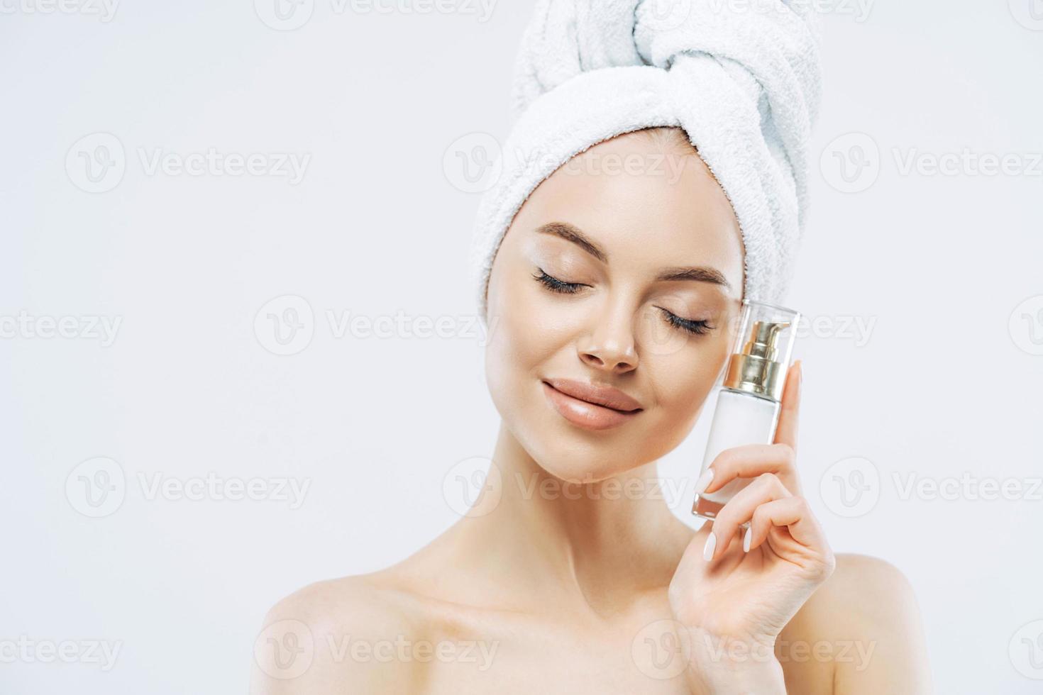 cuidado de la piel, concepto cosmético. foto de una joven europea relajada y sana parada con los ojos cerrados, sostiene una botella de producto cosmético para piel elástica, posa medio desnuda sobre fondo blanco.