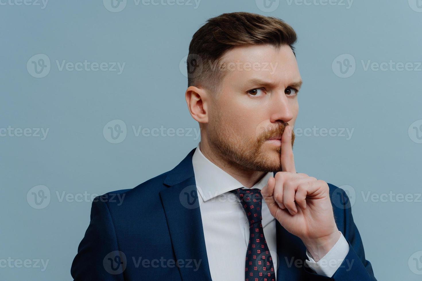 Serious bearded prosperous male office worker dressed in formal clothes makes hush gesture asks to keep quiet demands silence with strict expression isolated over blue background. Taboo shh shushing photo