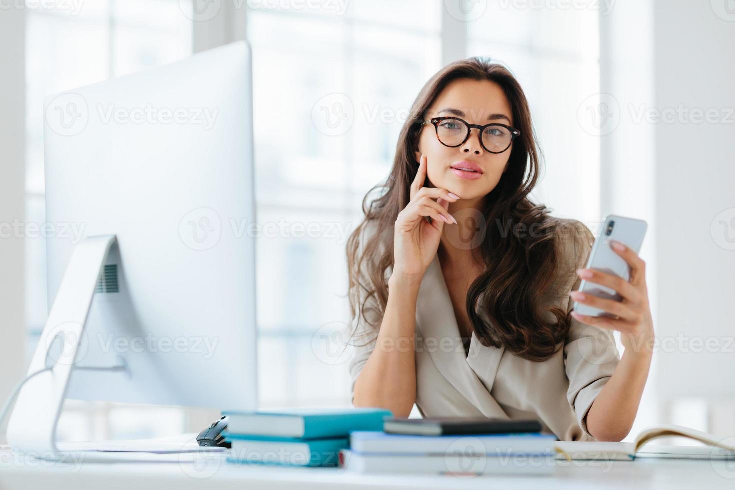 Female company office worker in formal wear uses modern gadget for work, poses at workplace near monitor, plans working schedule, browses information online, poses against window. Working time photo