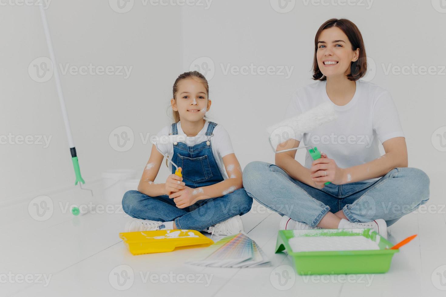 Photo of cheerful girl and daughter sit crossed legs on floor, hold paint rollers, paint room in white color, busy with refurbishment, have happy expressions. Home, repair, building and decoration