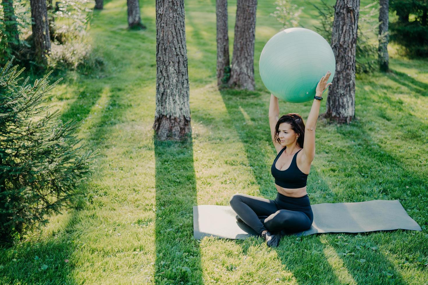 ejercicios de mujer deportiva en forma con pelota de fitness se sienta en karemat en pose de loto vestida con top recortado y polainas, posa en el bosque durante el día soleado, respira aire fresco. dama activa practica yoga afuera foto