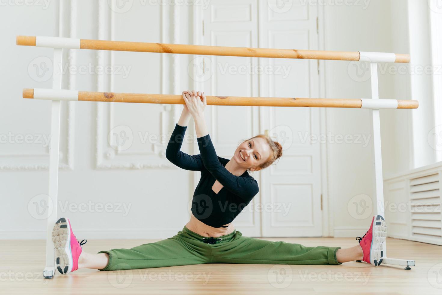 Caucasian female ballerina spreads legs sideways, shows splits, holds hands on barres, has ballet class, wears sportswear and sneakers, has perfect figure, tilts head on right, stretches herself. photo