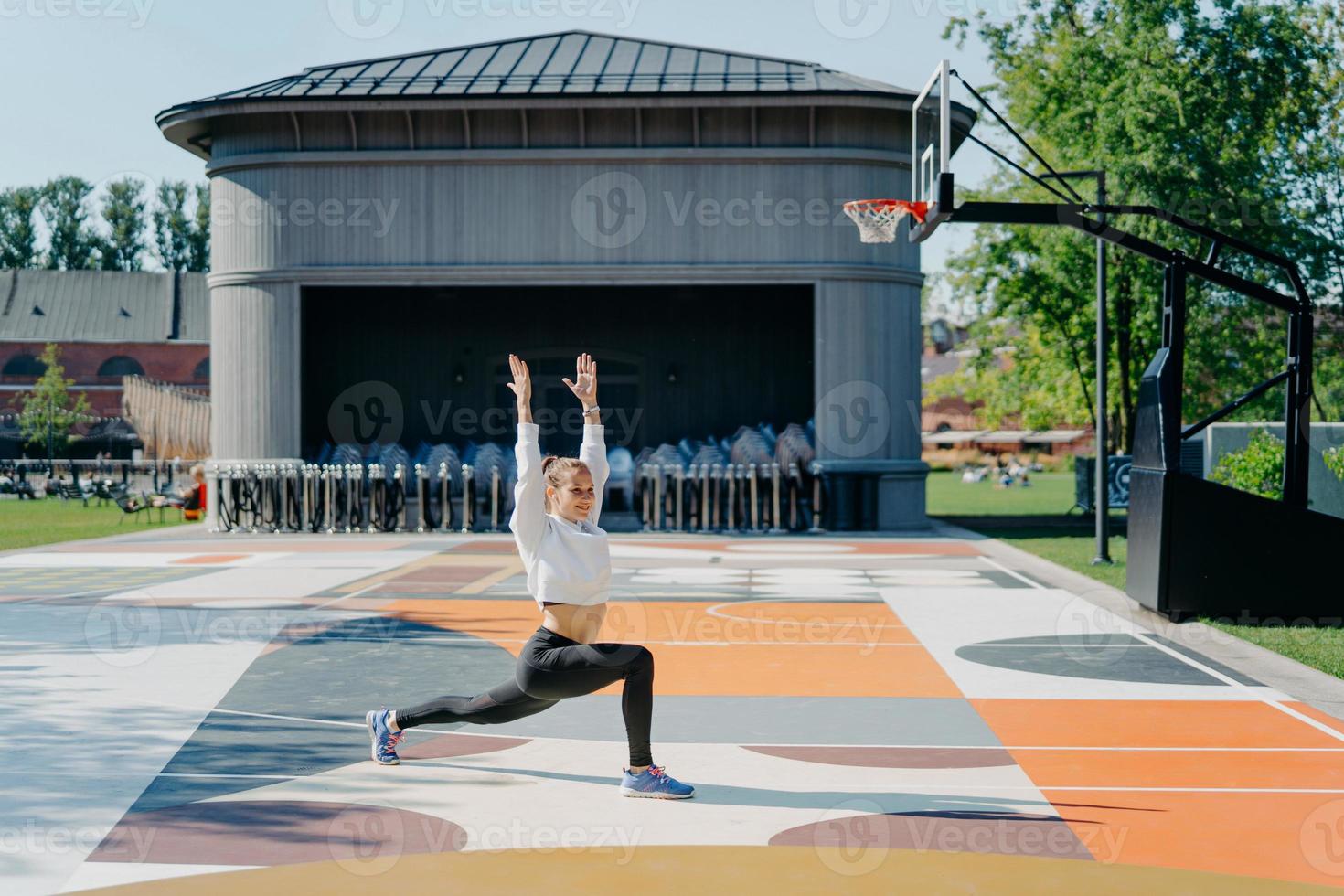 rutina de ejercicios personales. la mujer atlética hace ejercicios físicos al aire libre mantiene los brazos levantados se calienta antes de trotar vestida con poses de ropa activa en la cancha de baloncesto. flexibilidad y fitness foto