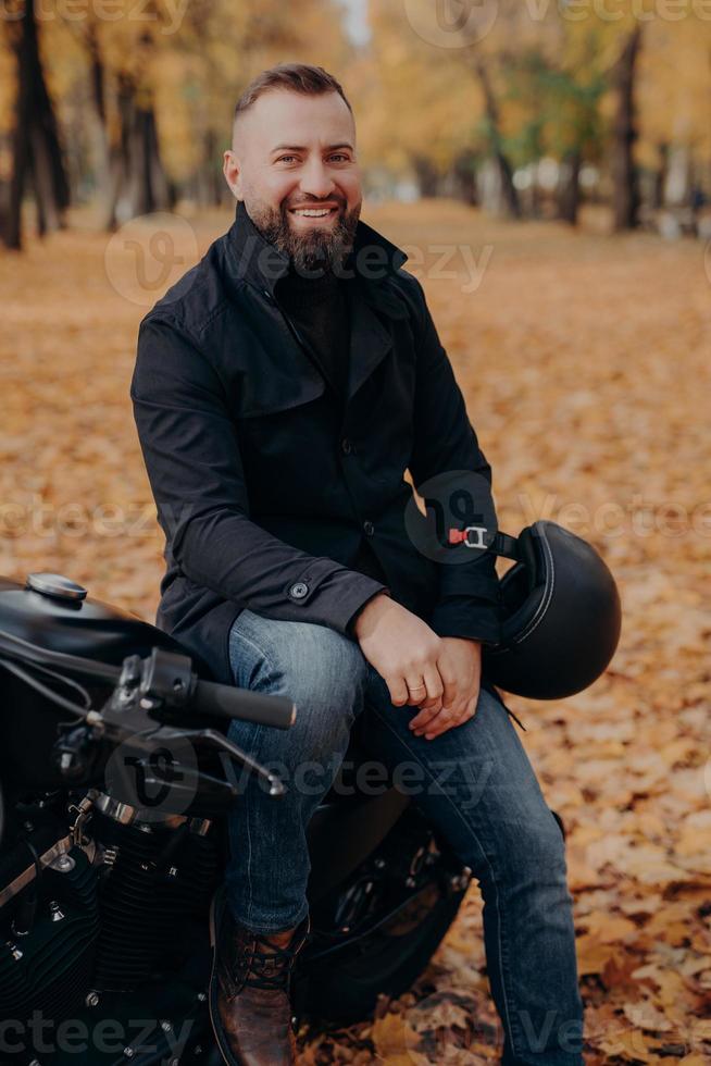 Vertical image of smiling motorcyclist smiles happily, holds helmet, poses on motorbike, wears black jacket and jeans, has drive through park, enjoys autumn time rides black vehicle. Transport concept photo