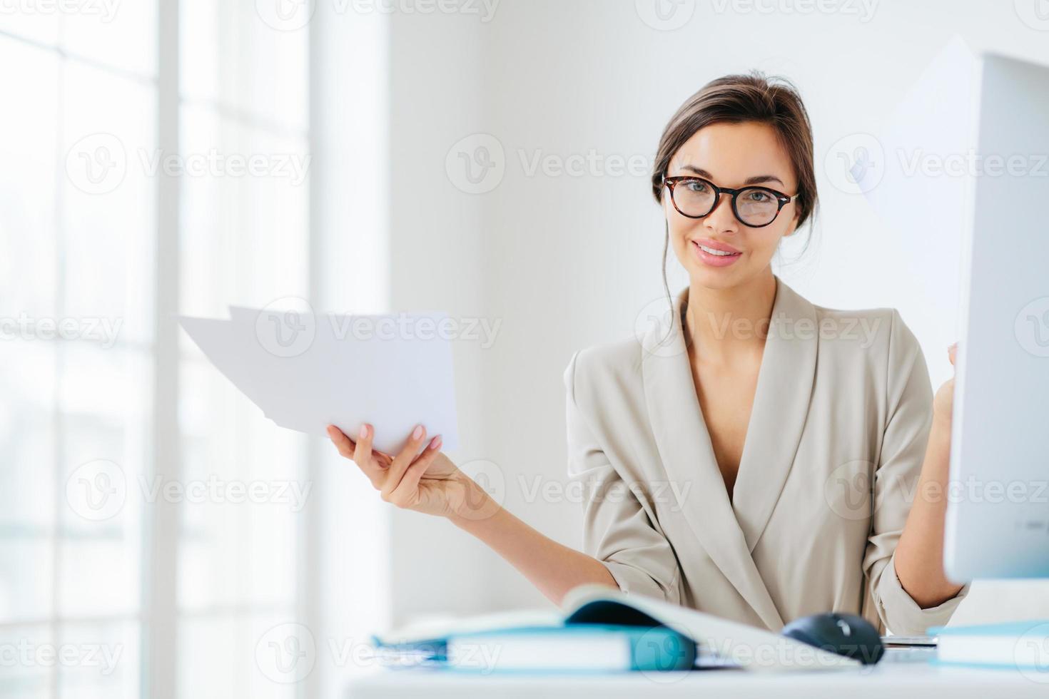 Indoor shot of good looking female in formal suit, holds paper documents, works on organization and creating startup project, poses at desktop with computer, checks information for presentation photo