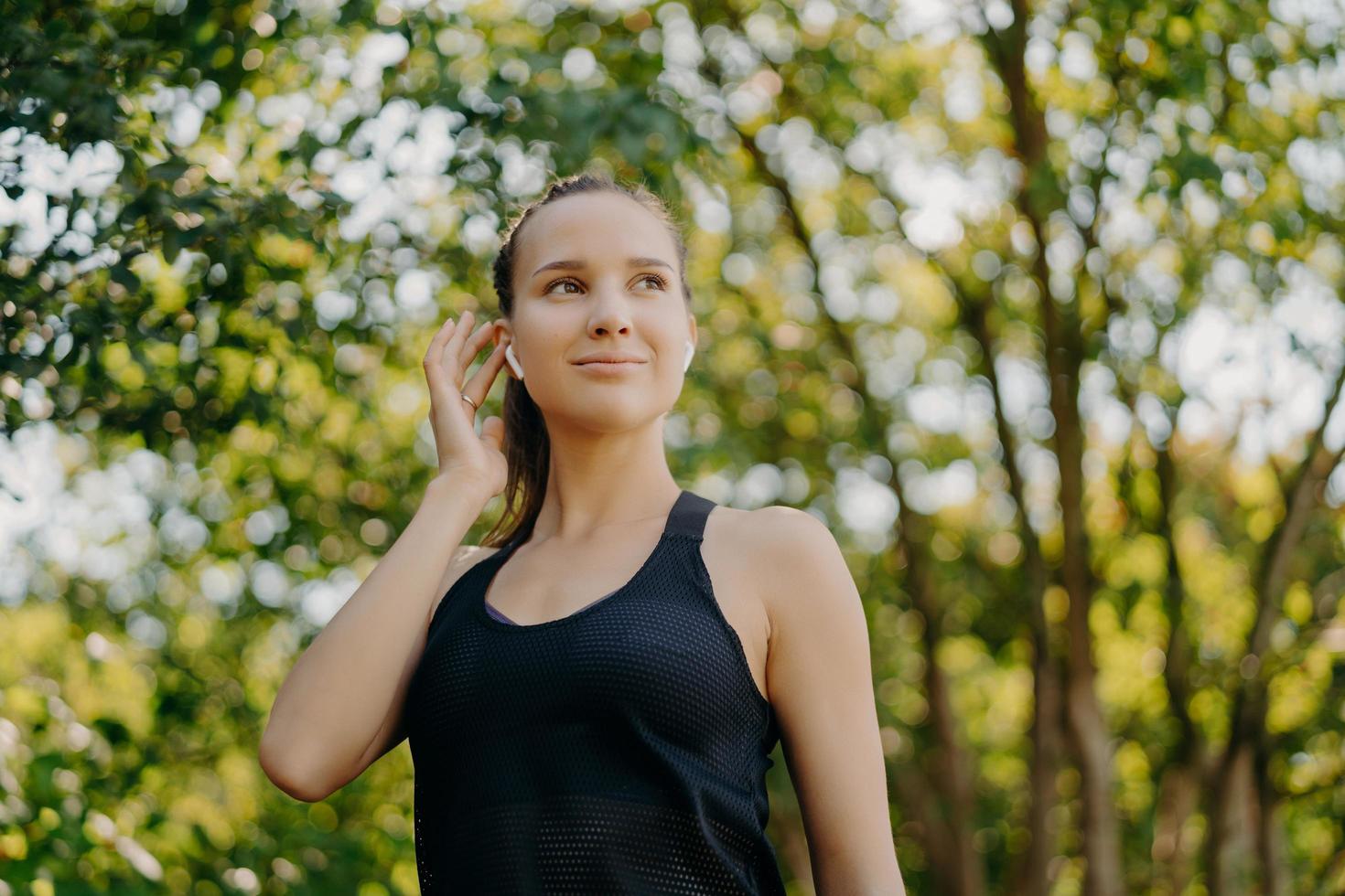 Horizontal shot of satisfied young woman keeps hand on earbud listens favorite music looks away has satisfied face expression dressed in active wear enjoys summer morning outdoors has cardio workout photo