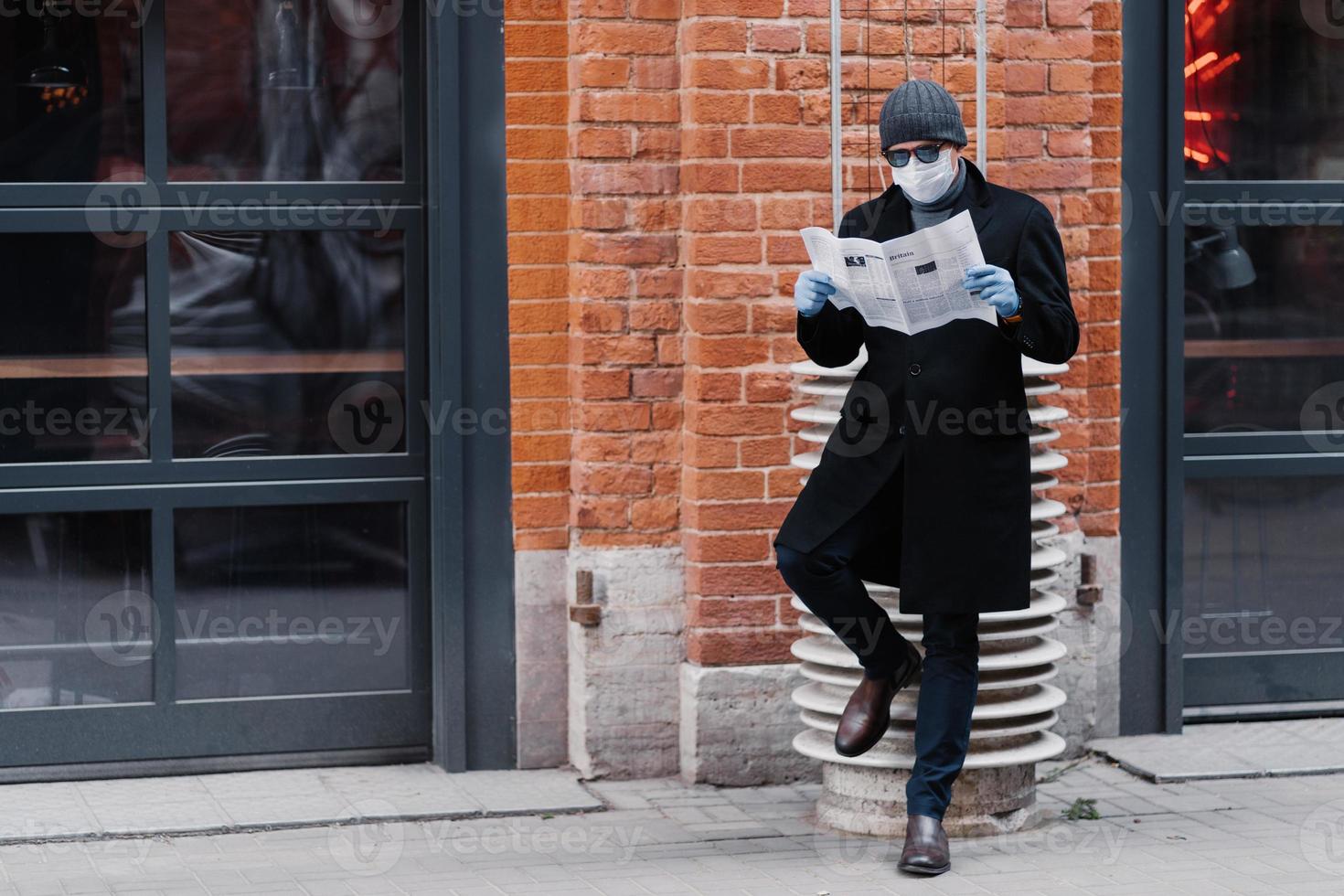 Full length shot of serious man dressed in black coat, wears sunglasses and medical mask, reads newspaper, poses against brick wall, prevents himself from coronavirus. Epidemic and quarantine photo