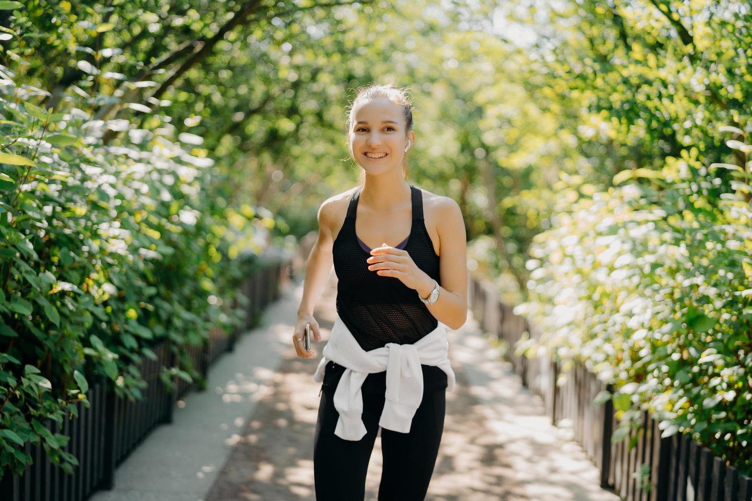 Sportive young woman has jogging workout training runs outdoor smiles pleasantly enjoys good day dressed in sportsclothes listens music from playlist. Start day from morning jog. Female runner photo