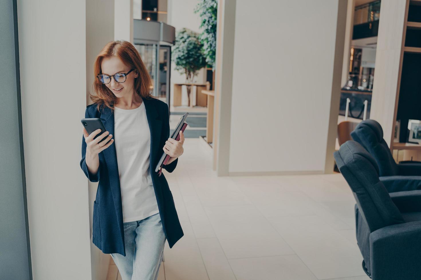 Smiling redhead female business consultant dressed formally holding mobile phone during coffee break photo