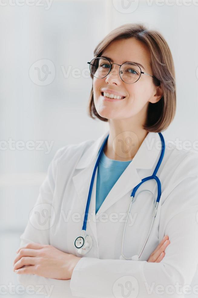 Close up portrait of short haired female general practitioner stands with smile and arms crossed, uses stethoscope, enjoys work, poses against white background. People, mediccare and treatment concept photo