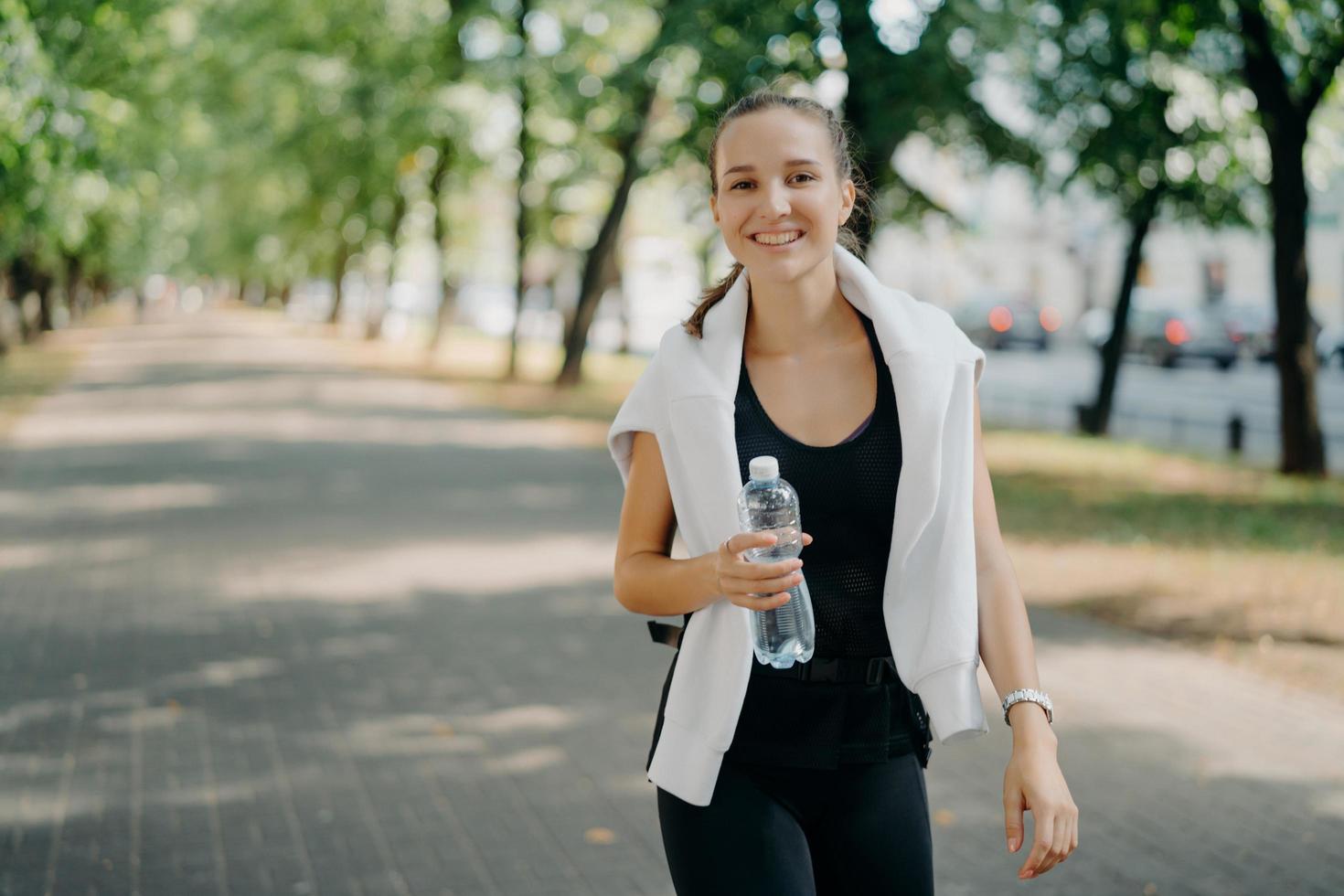 Outdoor shot of glad sporty woman drinks fresh water from bottle has walk during summer green urban park has toothy smile on face leads healthy lifestyle. Restoring aqua balanace after training photo